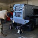 A military service member in MultiCam/OCP camouflage uniform is performing maintenance on a piece of ground support equipment labeled 'B807' inside a maintenance hangar or shop. The equipment appears to be a mobile generator or power unit mounted on a wheeled trailer, with jack stands placed underneath for support. The setting is an indoor military maintenance facility with tools and equipment visible in the background.