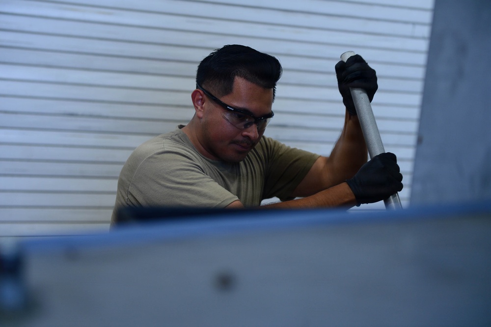 A man wearing safety glasses and black gloves is performing manual work using a metal rod or tool. He is dressed in an olive/tan t-shirt and appears to be working on or near a metal surface or equipment. The background shows corrugated metal siding, suggesting an industrial or outdoor work environment.