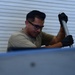 A man wearing safety glasses and black gloves is performing manual work using a metal rod or tool. He is dressed in an olive/tan t-shirt and appears to be working on or near a metal surface or equipment. The background shows corrugated metal siding, suggesting an industrial or outdoor work environment.