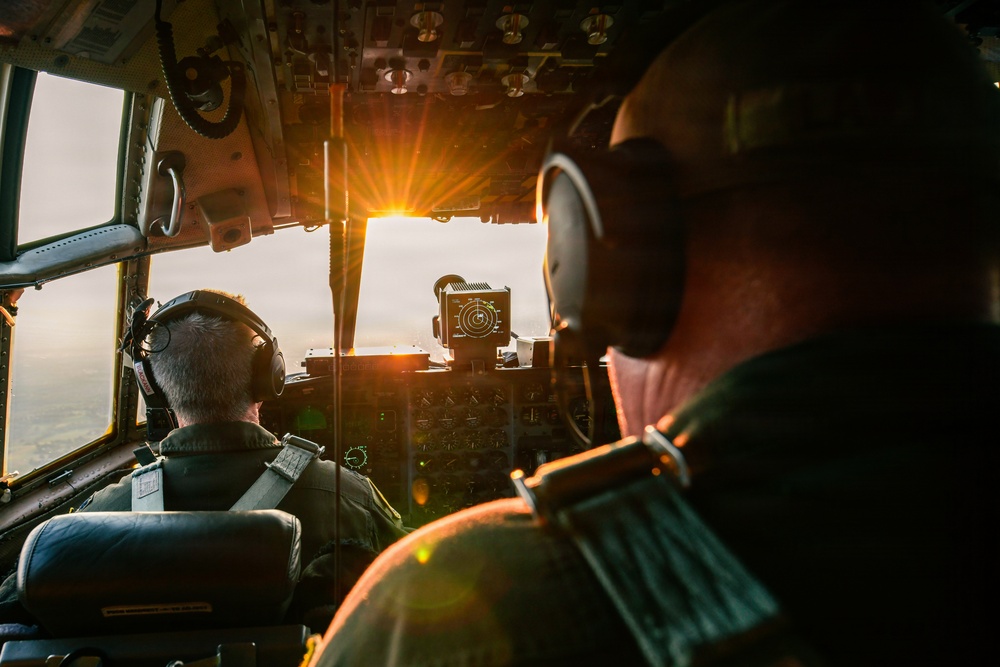 Two military aircrew members are seated in the cockpit of a large military aircraft, likely a C-130 Hercules or similar transport aircraft, during flight at either sunrise or sunset. The cockpit is bathed in warm golden light streaming through the windscreen, illuminating the instrument panels and crew. Both individuals are wearing flight helmets with integrated headsets and military flight suits.