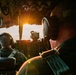 Two military aircrew members are seated in the cockpit of a large military aircraft, likely a C-130 Hercules or similar transport aircraft, during flight at either sunrise or sunset. The cockpit is bathed in warm golden light streaming through the windscreen, illuminating the instrument panels and crew. Both individuals are wearing flight helmets with integrated headsets and military flight suits.