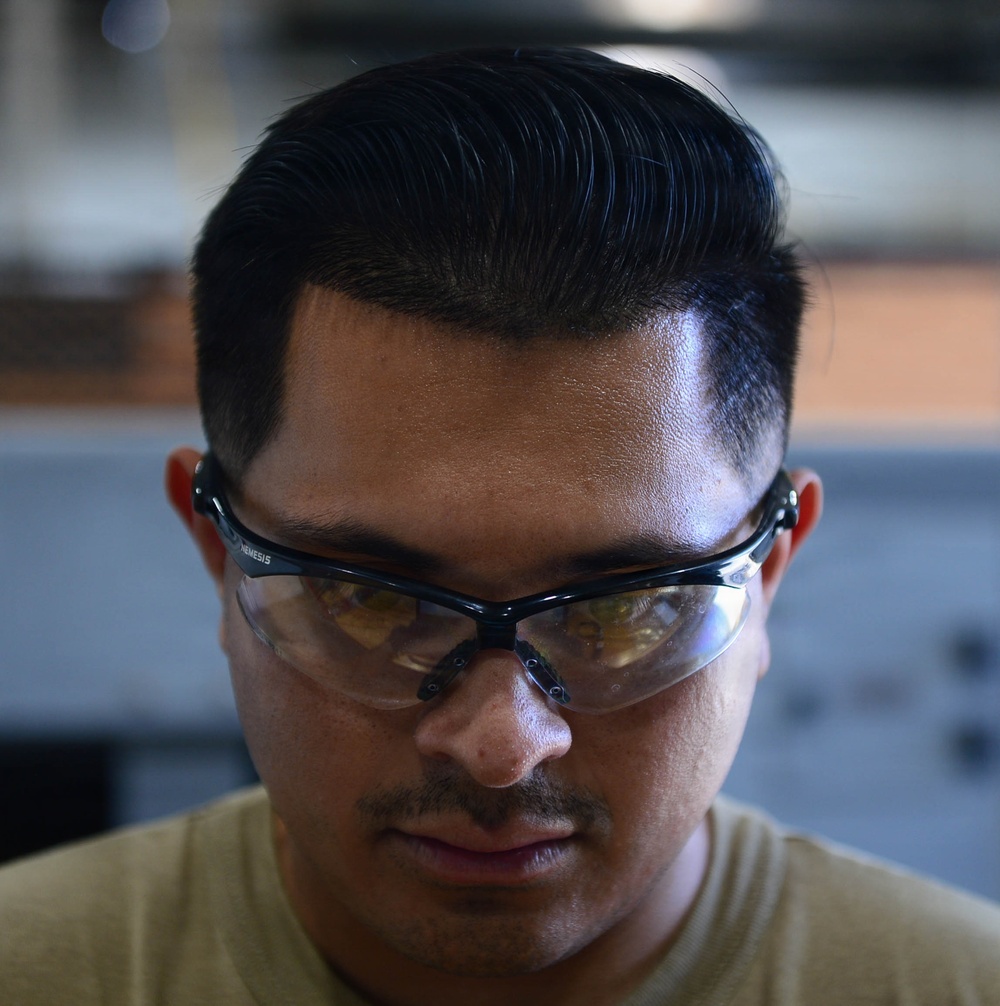 A close-up photograph of a young adult male wearing safety glasses, looking downward. He has a styled pompadour-fade haircut with dark hair and a light mustache/goatee. The background is blurred, suggesting an industrial or workshop environment.