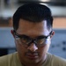 A close-up photograph of a young adult male wearing safety glasses, looking downward. He has a styled pompadour-fade haircut with dark hair and a light mustache/goatee. The background is blurred, suggesting an industrial or workshop environment.