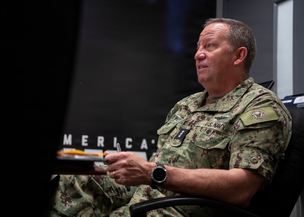 A U.S. Navy officer in NWU Type III camouflage uniform is seated in a chair, appearing to be engaged in a briefing or interview. The officer displays four-star rank insignia on the chest, indicating the rank of Admiral. A partial text reading 'MERICA' is visible in the background.