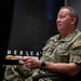 A U.S. Navy officer in NWU Type III camouflage uniform is seated in a chair, appearing to be engaged in a briefing or interview. The officer displays four-star rank insignia on the chest, indicating the rank of Admiral. A partial text reading 'MERICA' is visible in the background.