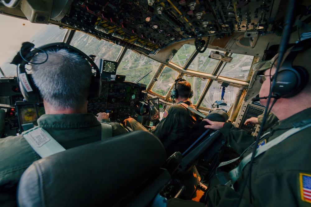 The image shows the interior cockpit of a large military transport aircraft, likely a C-130 Hercules, with three crew members visible from behind wearing olive drab flight suits and aviation headsets. The aircraft appears to be at low altitude over what looks like an airfield or urban area, visible through the multi-pane cockpit windows. The cockpit features an extensive array of analog and digital instruments, switches, and overhead panels typical of a military transport aircraft.