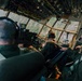 The image shows the interior cockpit of a large military transport aircraft, likely a C-130 Hercules, with three crew members visible from behind wearing olive drab flight suits and aviation headsets. The aircraft appears to be at low altitude over what looks like an airfield or urban area, visible through the multi-pane cockpit windows. The cockpit features an extensive array of analog and digital instruments, switches, and overhead panels typical of a military transport aircraft.
