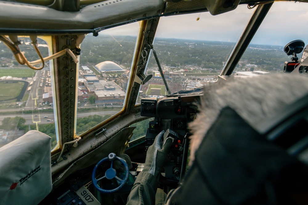 The image is taken from inside the cockpit of a vintage World War II-era bomber aircraft, likely a B-17 Flying Fortress or similar heavy bomber, in flight over a mid-sized American city. A pilot or crew member is visible operating the controls, with a yoke/control wheel and instrument panel visible in the foreground. Below, a football stadium, domed arena, and suburban landscape are clearly visible through the aged, riveted windscreen framing.