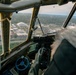The image is taken from inside the cockpit of a vintage World War II-era bomber aircraft, likely a B-17 Flying Fortress or similar heavy bomber, in flight over a mid-sized American city. A pilot or crew member is visible operating the controls, with a yoke/control wheel and instrument panel visible in the foreground. Below, a football stadium, domed arena, and suburban landscape are clearly visible through the aged, riveted windscreen framing.