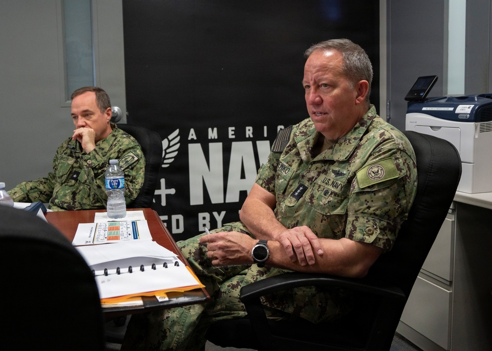 Two U.S. Navy personnel in digital camouflage (NWU Type III) uniforms are seated at a conference table, apparently in a briefing or meeting. The individual in the foreground appears to be a senior officer based on visible rank insignia (four stars visible on collar), and is speaking or presenting. An 'America's Navy' recruitment banner is visible on the wall behind them.