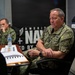Two U.S. Navy personnel in digital camouflage (NWU Type III) uniforms are seated at a conference table, apparently in a briefing or meeting. The individual in the foreground appears to be a senior officer based on visible rank insignia (four stars visible on collar), and is speaking or presenting. An 'America's Navy' recruitment banner is visible on the wall behind them.