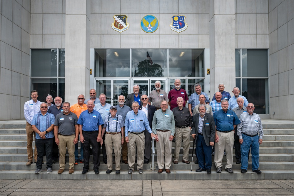 A large group of approximately 25-30 older men are posed for a formal group photograph on the steps of what appears to be a U.S. Air Force building. Three military insignia/shields are visible above the entrance doors, indicating an official military installation. The men are wearing civilian casual attire and most appear to have name tags, suggesting a reunion or commemorative event.
