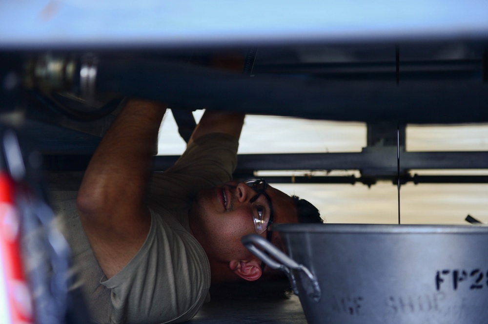 A technician or mechanic is lying on their back working underneath a large piece of equipment, likely an aircraft or military vehicle. The person is wearing glasses and a gray t-shirt, reaching upward to perform maintenance on components visible above them. The image is taken from a low angle, providing a close-up perspective of the work being performed.