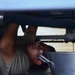 A technician or mechanic is lying on their back working underneath a large piece of equipment, likely an aircraft or military vehicle. The person is wearing glasses and a gray t-shirt, reaching upward to perform maintenance on components visible above them. The image is taken from a low angle, providing a close-up perspective of the work being performed.
