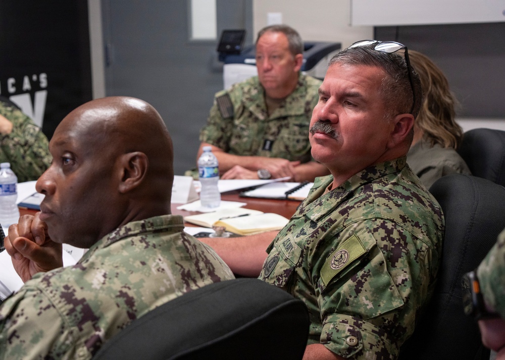 A group of military personnel in camouflage uniforms are seated around a conference table, appearing to attend a briefing or meeting. At least four individuals are visible, wearing what appears to be US Navy Working Uniform (NWU) digital camouflage pattern. The setting is an indoor conference room with documents, notebooks, and water bottles on the table.