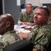 A group of military personnel in camouflage uniforms are seated around a conference table, appearing to attend a briefing or meeting. At least four individuals are visible, wearing what appears to be US Navy Working Uniform (NWU) digital camouflage pattern. The setting is an indoor conference room with documents, notebooks, and water bottles on the table.