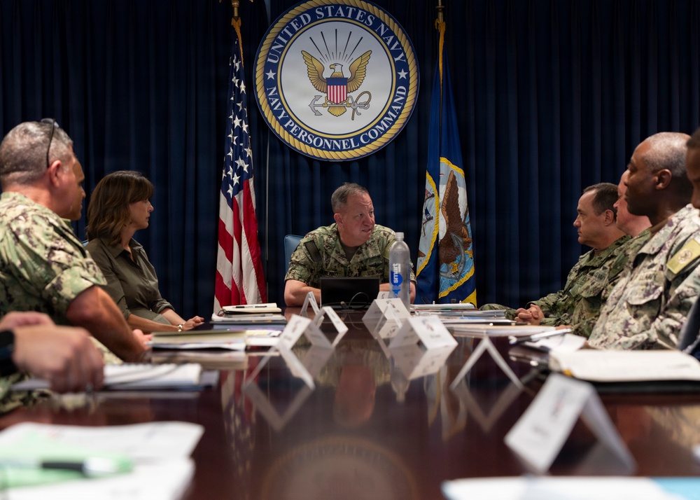 A formal military briefing or meeting is taking place at Navy Personnel Command headquarters. Several U.S. Navy personnel in uniform, along with at least one civilian, are seated around a large conference table. The central figure appears to be presiding over the meeting, with the Navy Personnel Command seal prominently displayed on the wall behind him.
