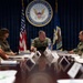 A formal military briefing or meeting is taking place at Navy Personnel Command headquarters. Several U.S. Navy personnel in uniform, along with at least one civilian, are seated around a large conference table. The central figure appears to be presiding over the meeting, with the Navy Personnel Command seal prominently displayed on the wall behind him.