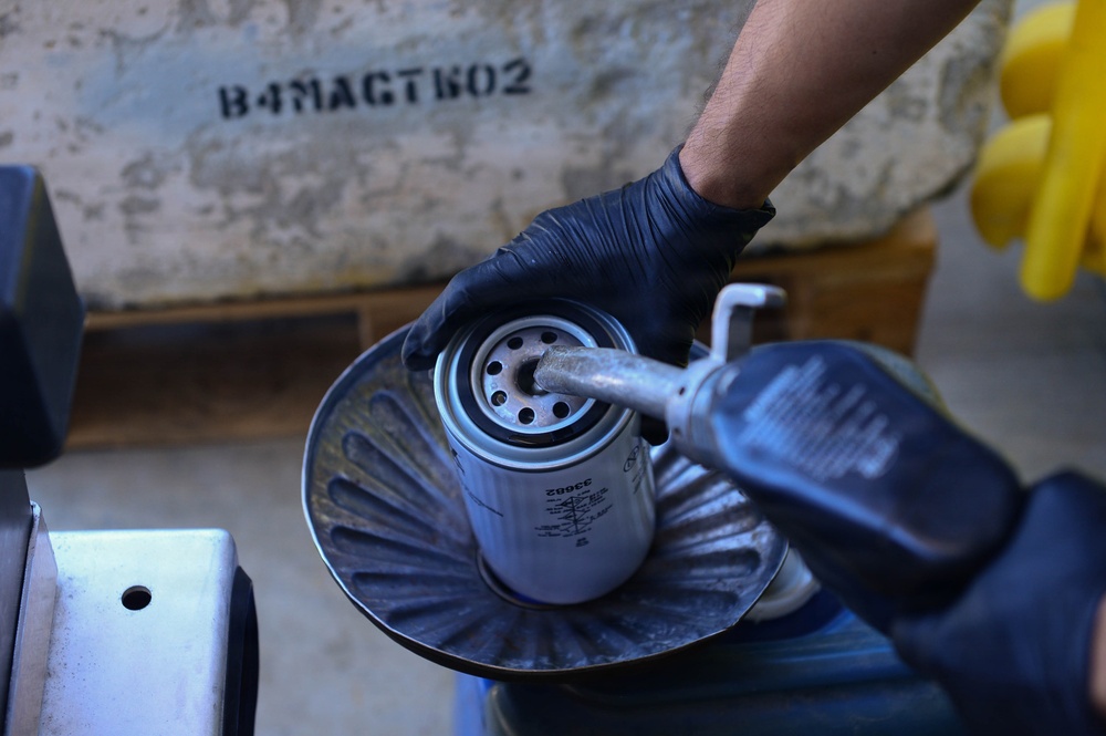A mechanic wearing black nitrile gloves is removing or handling an oil filter using a filter wrench tool, holding it over a metal drain pan to catch any residual oil. The setting appears to be an automotive repair garage or workshop. A stenciled text 'В4МАСТКО2' is visible on the concrete wall in the background.