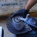 A mechanic wearing black nitrile gloves is removing or handling an oil filter using a filter wrench tool, holding it over a metal drain pan to catch any residual oil. The setting appears to be an automotive repair garage or workshop. A stenciled text 'В4МАСТКО2' is visible on the concrete wall in the background.