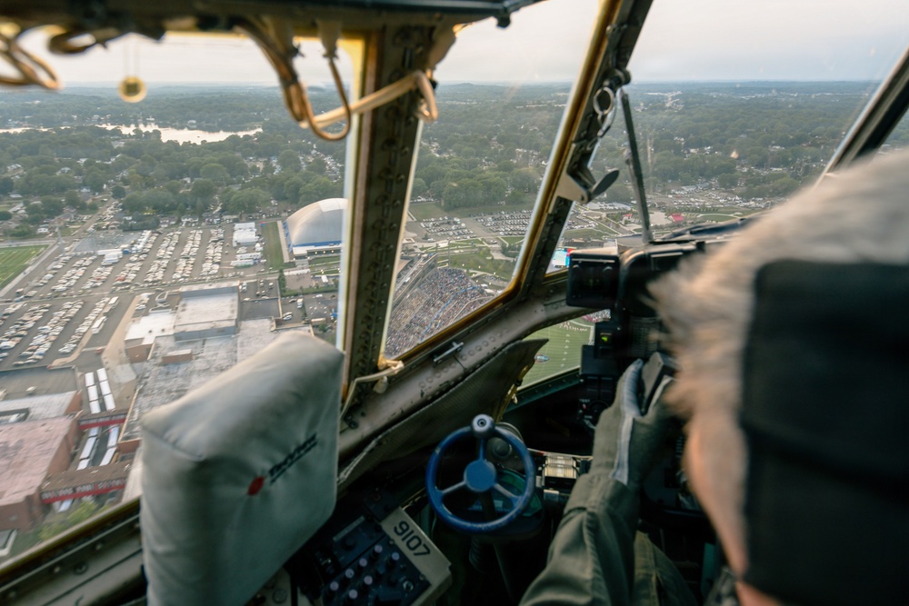 The image is taken from inside the cockpit of a vintage military aircraft, likely a B-17 or similar WWII-era bomber, during a low-altitude flyover of a stadium and surrounding area. A pilot or crew member is visible on the right side operating the aircraft controls, including a distinctive blue steering yoke. Below, a football stadium filled with spectators, parking lots, and a domed building are clearly visible.
