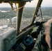 The image is taken from inside the cockpit of a vintage military aircraft, likely a B-17 or similar WWII-era bomber, during a low-altitude flyover of a stadium and surrounding area. A pilot or crew member is visible on the right side operating the aircraft controls, including a distinctive blue steering yoke. Below, a football stadium filled with spectators, parking lots, and a domed building are clearly visible.