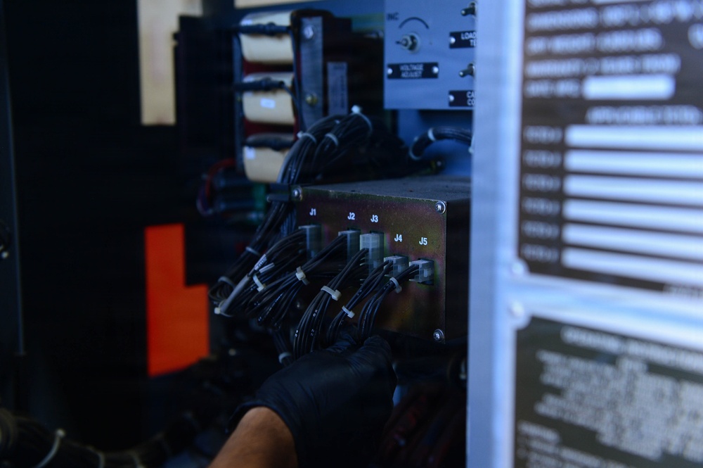 A technician wearing black nitrile gloves is working on an electrical or electronic equipment panel, handling multiple bundled black cables connected to labeled ports. The panel features connector ports labeled J1 through J5, with numerous cable harnesses routed through the assembly. The equipment appears to be a military or industrial-grade power or control unit based on the visible warning placards and hardware construction.