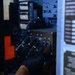 A technician wearing black nitrile gloves is working on an electrical or electronic equipment panel, handling multiple bundled black cables connected to labeled ports. The panel features connector ports labeled J1 through J5, with numerous cable harnesses routed through the assembly. The equipment appears to be a military or industrial-grade power or control unit based on the visible warning placards and hardware construction.