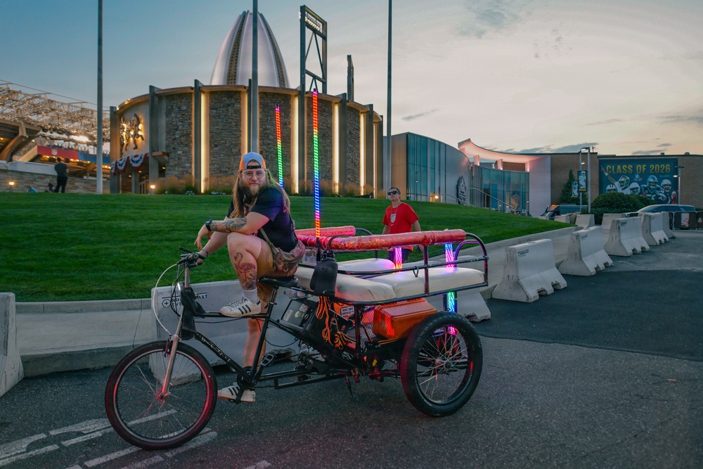 A tattooed man with long hair and a backwards cap sits on a decorated pedicab/rickshaw tricycle adorned with colorful LED lights near dusk. The setting appears to be outside the Pro Football Hall of Fame in Canton, Ohio, evidenced by the distinctive rotunda building and a 'Class of 2026' banner visible in the background. The scene captures a festive atmosphere likely tied to an NFL Hall of Fame induction event.