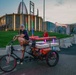 A tattooed man with long hair and a backwards cap sits on a decorated pedicab/rickshaw tricycle adorned with colorful LED lights near dusk. The setting appears to be outside the Pro Football Hall of Fame in Canton, Ohio, evidenced by the distinctive rotunda building and a 'Class of 2026' banner visible in the background. The scene captures a festive atmosphere likely tied to an NFL Hall of Fame induction event.