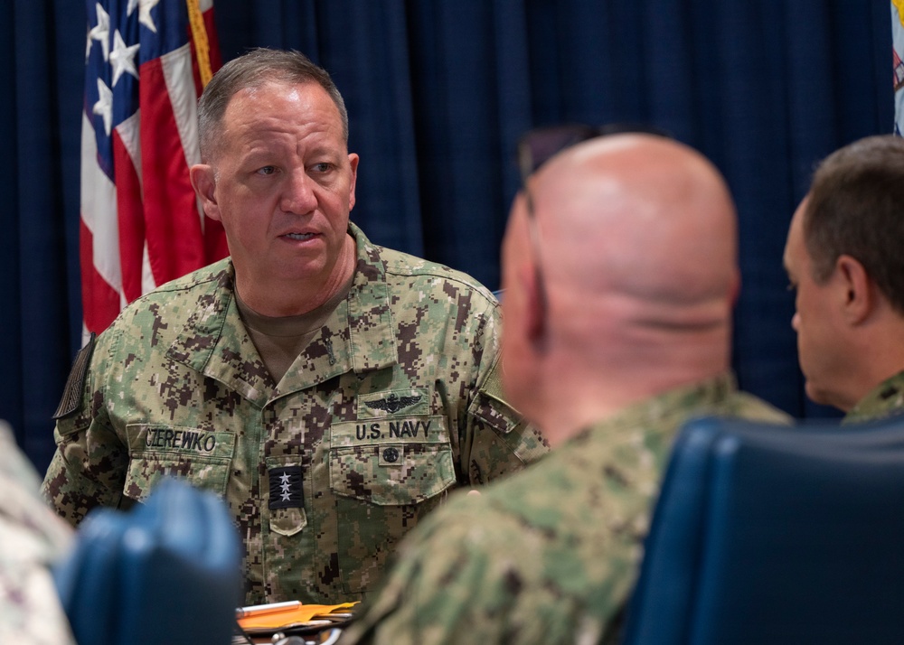 A U.S. Navy officer with the name tape 'CIEREWKO' and rank insignia of a Vice Admiral (three stars) is engaged in conversation with other military personnel at what appears to be a formal meeting or briefing. He is wearing the Navy Working Uniform (NWU Type III) with a Naval Aviator badge and 'U.S. NAVY' tape visible. An American flag and what appears to be a command flag are visible in the background.