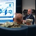 A civilian presenter in a dark suit is briefing military personnel on the 'Navy Personnel Data Ecosystem,' as shown on a large display screen behind him. Two uniformed Navy personnel are seated in the foreground with their backs to the camera, listening to the presentation. The slide details data architecture components including a Hard Pay Boundary, Pay System, Jupiter/Advana digital twin updates, and Authoritative Personnel Data Layer.
