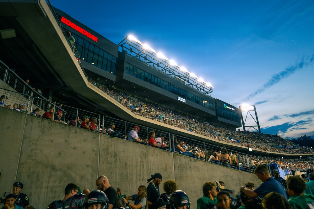 A nighttime sporting event at a large multi-tiered stadium filled with spectators. The photo is taken from field level looking up at the stands, with stadium lights illuminating the scene against a deep blue dusk sky. Visible sponsor signage includes 'Hendrickson' and 'Cleveland Clinic', and players or staff in helmets and uniforms are visible in the foreground.
