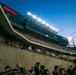 A nighttime sporting event at a large multi-tiered stadium filled with spectators. The photo is taken from field level looking up at the stands, with stadium lights illuminating the scene against a deep blue dusk sky. Visible sponsor signage includes 'Hendrickson' and 'Cleveland Clinic', and players or staff in helmets and uniforms are visible in the foreground.