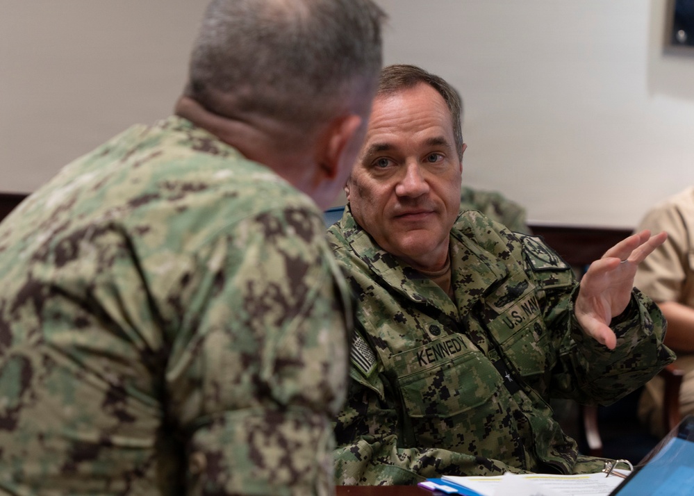 Two U.S. Navy personnel in Navy Working Uniform (NWU) camouflage are engaged in a discussion at what appears to be a meeting or briefing table. The individual facing the camera has a name tape reading 'KENNEDY' and a 'U.S. NAVY' tape visible on their uniform. The setting appears to be an indoor military facility with documents and binders on the table.