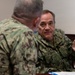 Two U.S. Navy personnel in Navy Working Uniform (NWU) camouflage are engaged in a discussion at what appears to be a meeting or briefing table. The individual facing the camera has a name tape reading 'KENNEDY' and a 'U.S. NAVY' tape visible on their uniform. The setting appears to be an indoor military facility with documents and binders on the table.