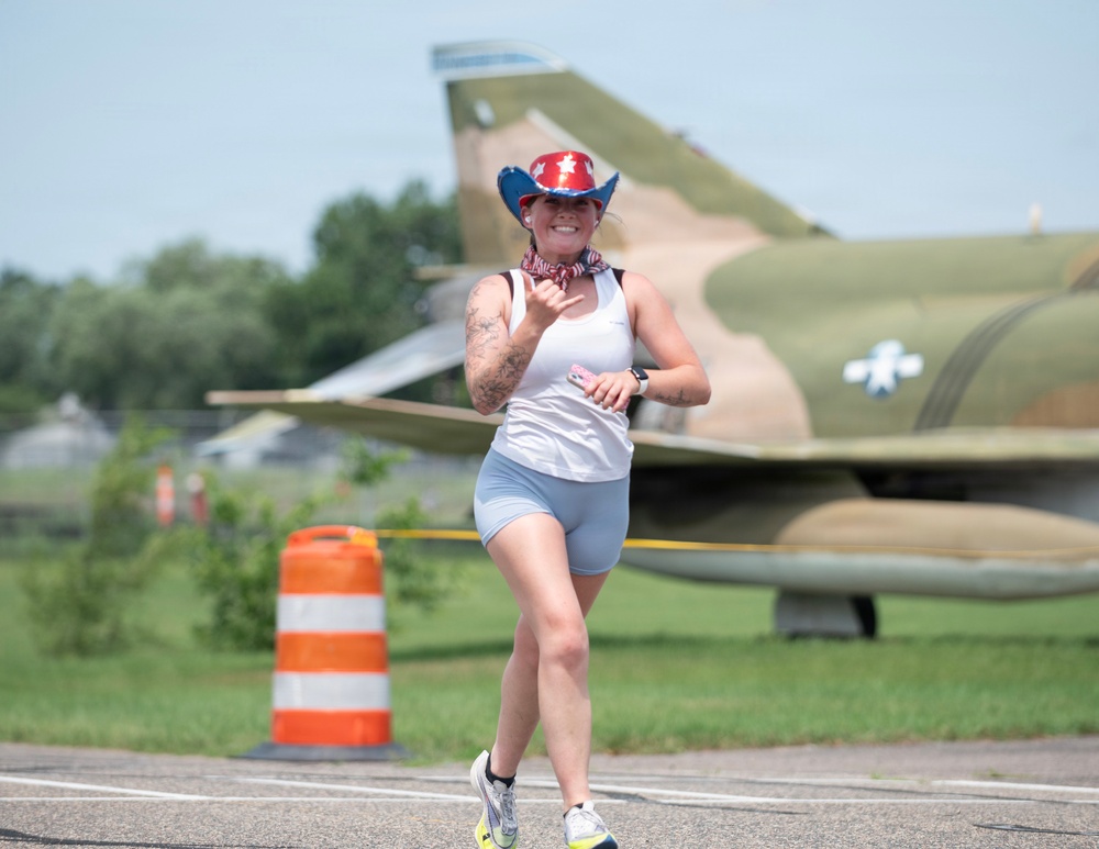 A woman is running on a paved surface near a military airfield, wearing patriotic-themed accessories including a red, white, and blue cowboy hat and a bandana. She is smiling and making a 'shaka' hand gesture toward the camera. A camouflaged military jet aircraft with a US Air Force roundel is visible in the background.