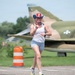 A woman is running on a paved surface near a military airfield, wearing patriotic-themed accessories including a red, white, and blue cowboy hat and a bandana. She is smiling and making a 'shaka' hand gesture toward the camera. A camouflaged military jet aircraft with a US Air Force roundel is visible in the background.