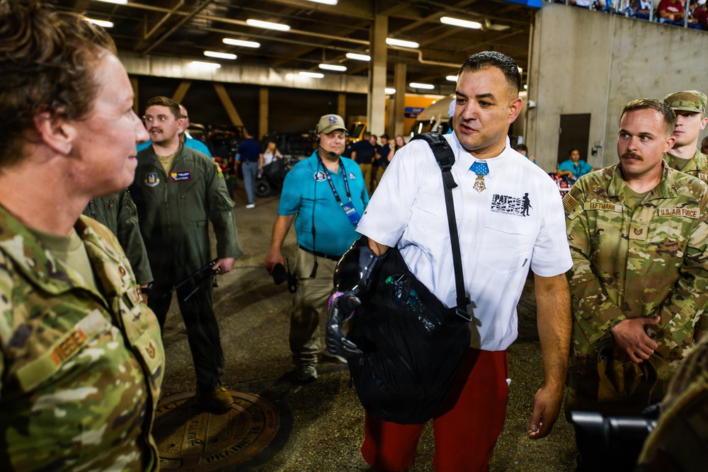 A group of people are gathered in what appears to be a stadium concourse or tunnel area. A civilian man wearing a white shirt with a Medal of Honor replica and red shorts is conversing with U.S. Air Force personnel in camouflage uniforms. The setting suggests a military appreciation or patriotic event at a sports venue.
