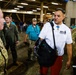A group of people are gathered in what appears to be a stadium concourse or tunnel area. A civilian man wearing a white shirt with a Medal of Honor replica and red shorts is conversing with U.S. Air Force personnel in camouflage uniforms. The setting suggests a military appreciation or patriotic event at a sports venue.