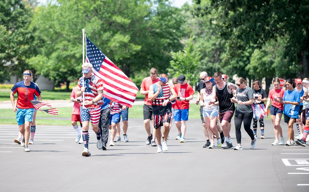 A large group of people are participating in an outdoor patriotic-themed run or race event, likely on or around the Fourth of July. A central participant is prominently carrying a large American flag on a pole while running. The majority of participants are dressed in red, white, and blue attire with American flag patterns.