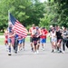 A large group of people are participating in an outdoor patriotic-themed run or race event, likely on or around the Fourth of July. A central participant is prominently carrying a large American flag on a pole while running. The majority of participants are dressed in red, white, and blue attire with American flag patterns.