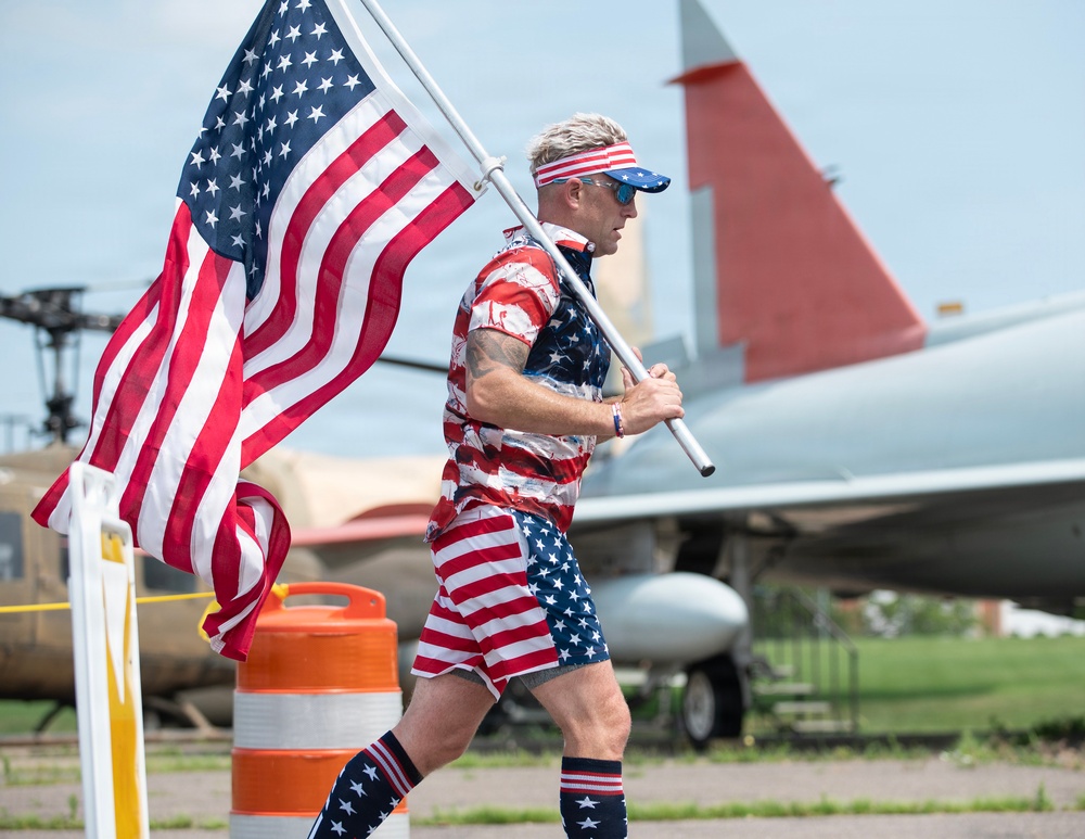 A man dressed entirely in American flag-themed clothing is running or walking while carrying a large United States flag on a pole. The setting appears to be near a military aviation museum or airfield, with vintage aircraft visible in the background. The patriotic outfit and flag suggest this may be a patriotic event, race, or holiday celebration.