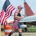 A man dressed entirely in American flag-themed clothing is running or walking while carrying a large United States flag on a pole. The setting appears to be near a military aviation museum or airfield, with vintage aircraft visible in the background. The patriotic outfit and flag suggest this may be a patriotic event, race, or holiday celebration.