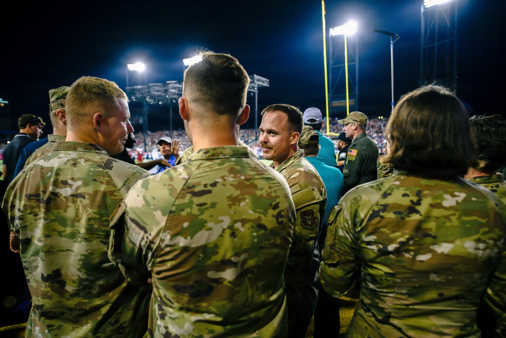 A group of military personnel in OCP (Operational Camouflage Pattern) uniforms are gathered on the sideline of an American football stadium during a night game. The individuals appear to be participating in a military appreciation or recognition event at the stadium. Stadium lights, goal posts, and a crowd of spectators are visible in the background.