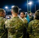 A group of military personnel in OCP (Operational Camouflage Pattern) uniforms are gathered on the sideline of an American football stadium during a night game. The individuals appear to be participating in a military appreciation or recognition event at the stadium. Stadium lights, goal posts, and a crowd of spectators are visible in the background.