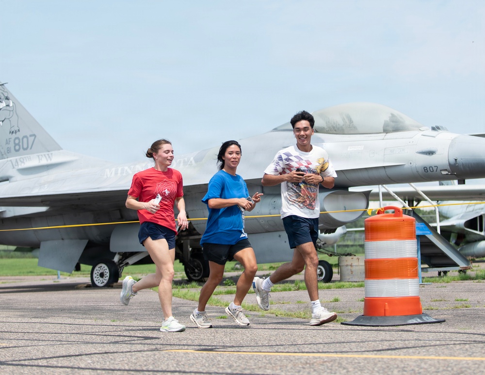Three individuals are jogging on a military airfield tarmac past a parked F-16 Fighting Falcon jet aircraft. The aircraft bears the markings 'AF 81 807', '148th FW', and 'MINNESOTA', identifying it as belonging to the 148th Fighter Wing of the Minnesota Air National Guard. An orange traffic barrel is visible in the foreground, suggesting a marked running course or safety boundary.