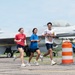 Three individuals are jogging on a military airfield tarmac past a parked F-16 Fighting Falcon jet aircraft. The aircraft bears the markings 'AF 81 807', '148th FW', and 'MINNESOTA', identifying it as belonging to the 148th Fighter Wing of the Minnesota Air National Guard. An orange traffic barrel is visible in the foreground, suggesting a marked running course or safety boundary.