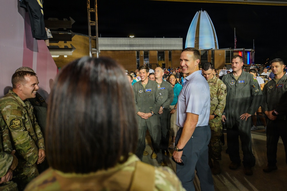 A civilian man in a light polo shirt and gray pants is interacting with a group of military personnel, including individuals in flight suits and camouflage uniforms, at what appears to be an outdoor nighttime event. The gathering has a celebratory or meet-and-greet atmosphere, with personnel smiling and engaged in conversation. A distinctive illuminated spire-like structure is visible in the background.
