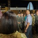 A civilian man in a light polo shirt and gray pants is interacting with a group of military personnel, including individuals in flight suits and camouflage uniforms, at what appears to be an outdoor nighttime event. The gathering has a celebratory or meet-and-greet atmosphere, with personnel smiling and engaged in conversation. A distinctive illuminated spire-like structure is visible in the background.